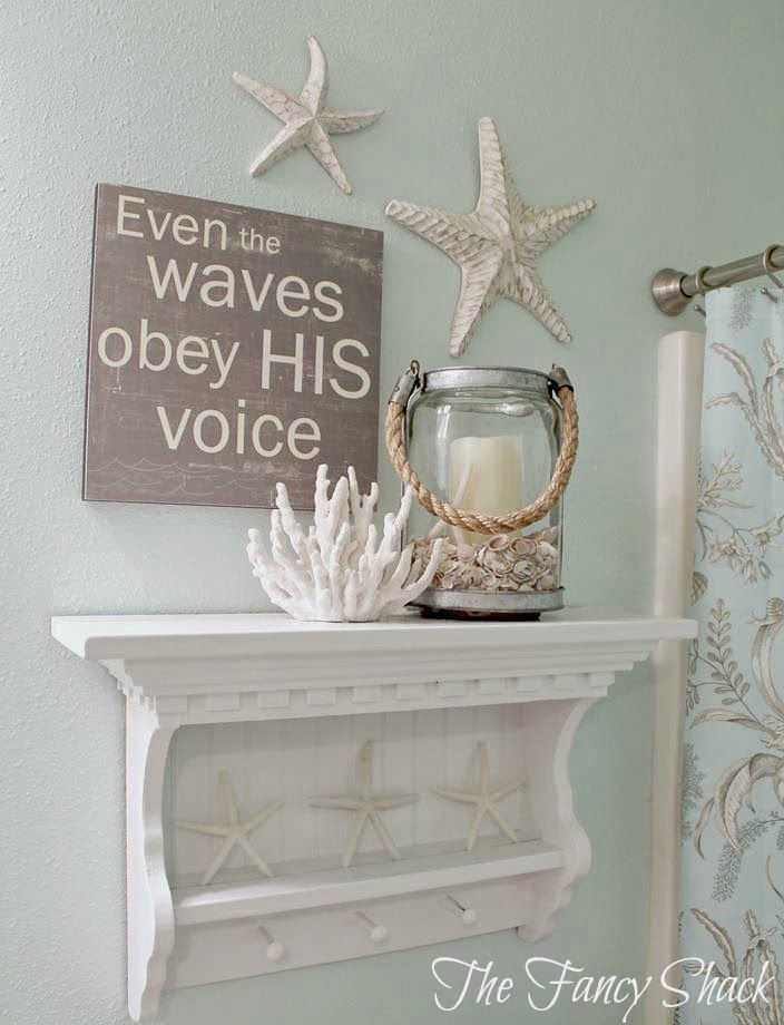 This charming bathroom display combines faith and coastal elements, featuring a sign with an inspirational message alongside starfish decorations. A white shelf adorned with coral and a candle lantern adds to the beachy, serene atmosphere.